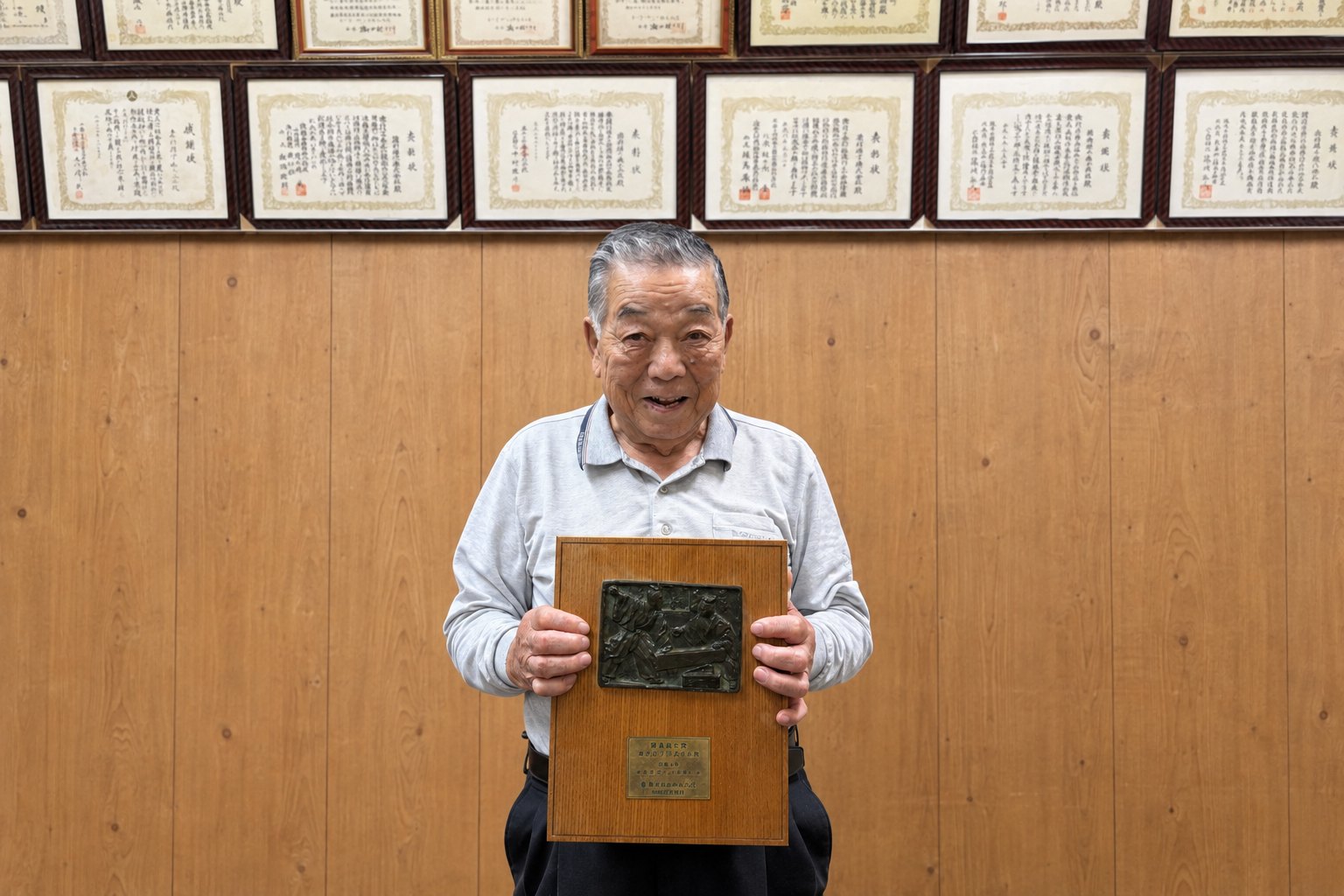 表彰状が並ぶ事務所の壁の前で、清水建設株式会社の優良施工賞の盾を持つ会長 霜村 研一